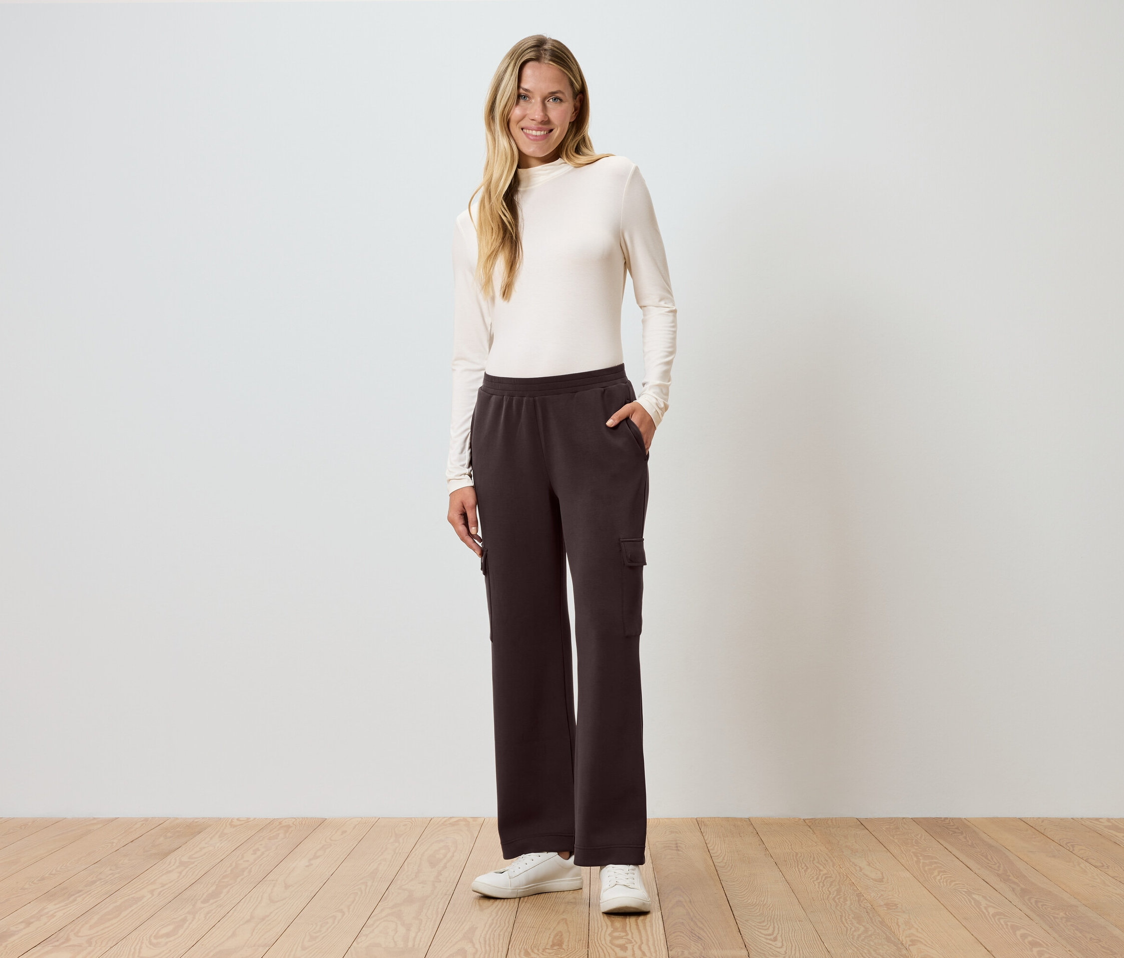 Kobieta stoi z ręką w kieszeni przed ścianą, ubrana w brązowe spodnie cargo.