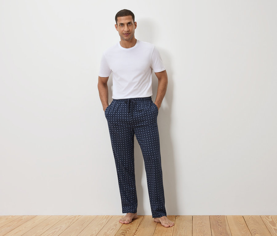 Mężczyzna stoi na tle białej ściany, ubrany w biały T-shirt i granatowe spodnie od piżamy w kropki.