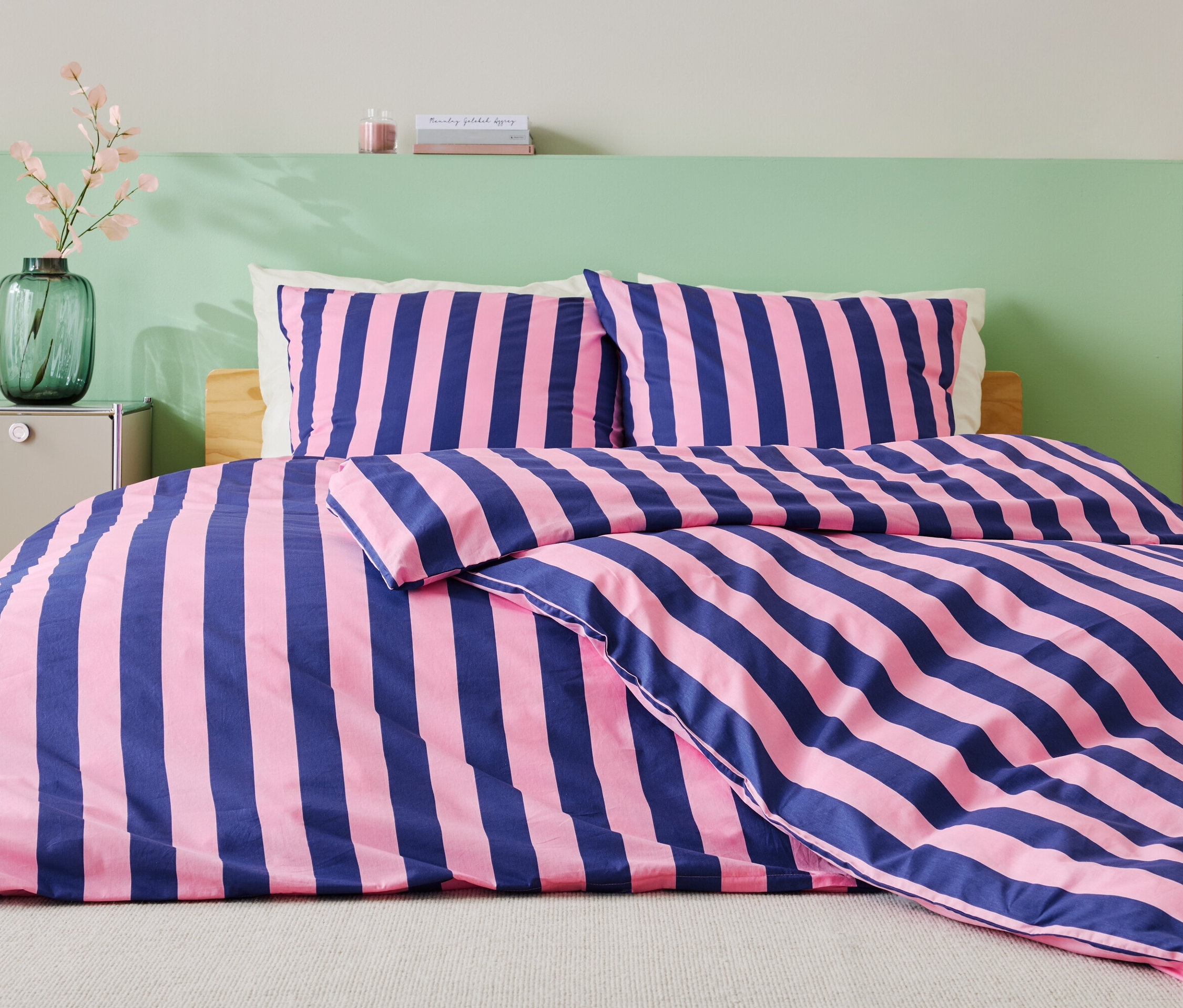 Łóżko z niebiesko-różową pościelą w paski, dwiema poduszkami i zieloną ścianą w tle.