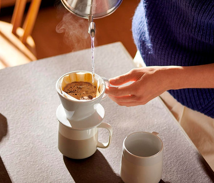 Osoba nalewa gorącą wodę z czajnika do białego ręcznego filtra do kawy, rozm. 101, umieszczonego na kubku. Z filtra unosi się para. Obok na stole stoi drugi kubek.