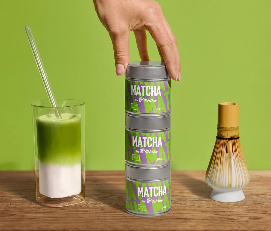 Ręka układa trzy puszki Bio Matcha by Tchibo – 30 g. Matcha latte w szklance ze słomką i miotełka do matchy na drewnie przed zielenią.