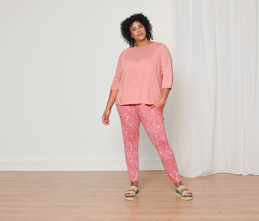 Kobieta pozuje w koralowej bluzce z rękawami o długości 3/4, koralowych spodniach dżersejowych z nadrukiem na całej powierzchni i skórzanych sandałach na platformie.