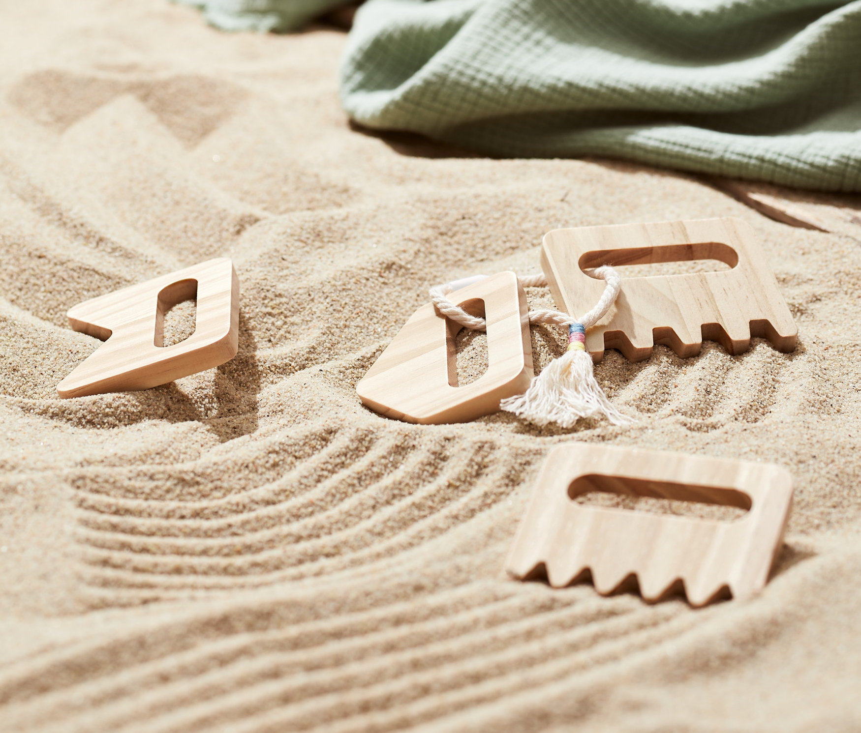 Na piasku leżą trzy drewniane grabki do piasku, zielony koc i japonki.