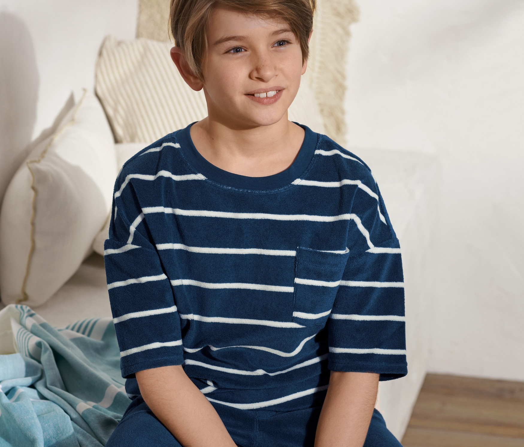 Chłopiec siedzi na łóżku i ma na sobie koszulkę z tkaniny frotte w niebiesko-białe paski.