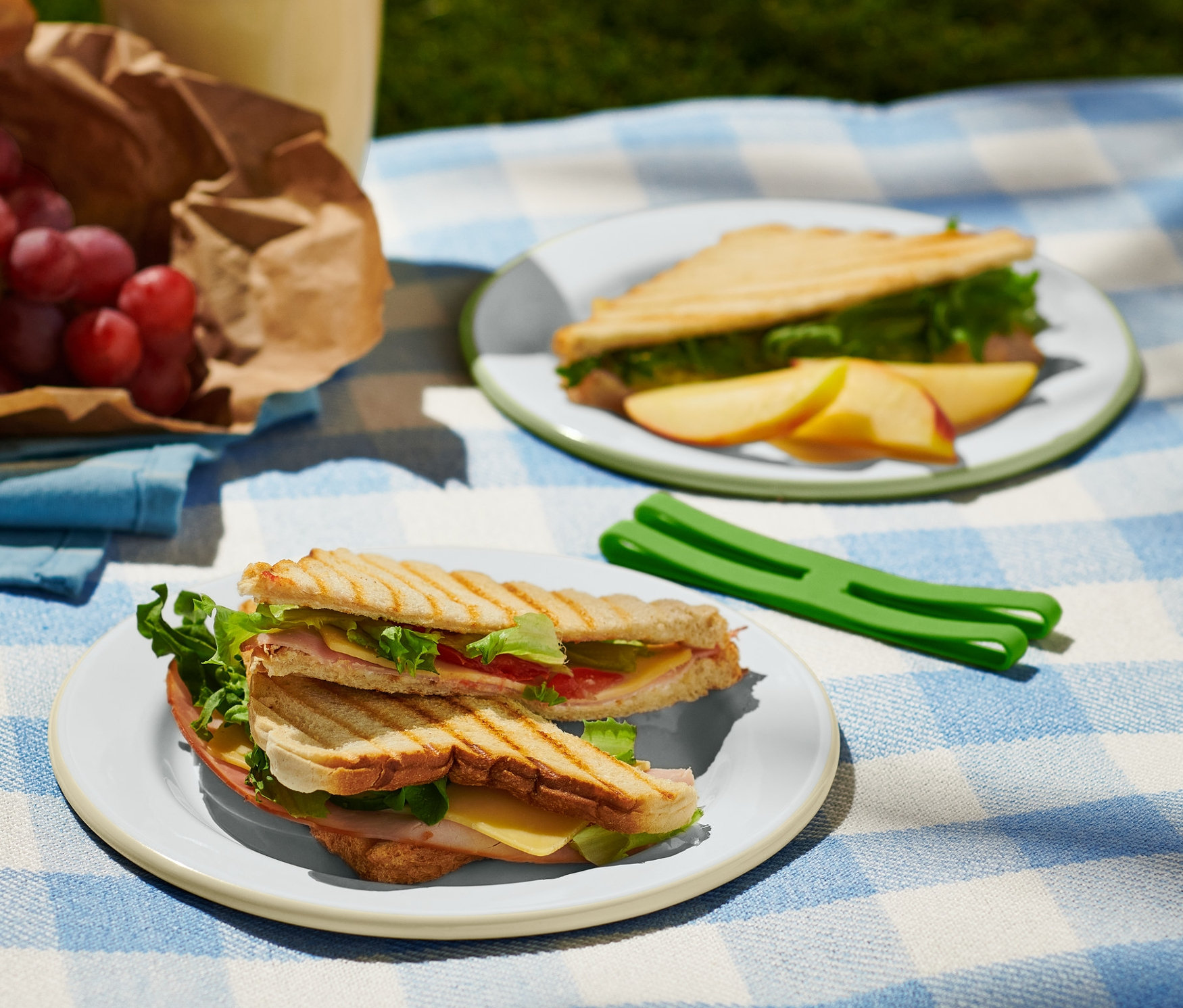 Na niebiesko-białym kocu w kratę leżą dwa talerze piknikowe, 2 sztuki z tostami, winogrona w papierowej torbie i napój.