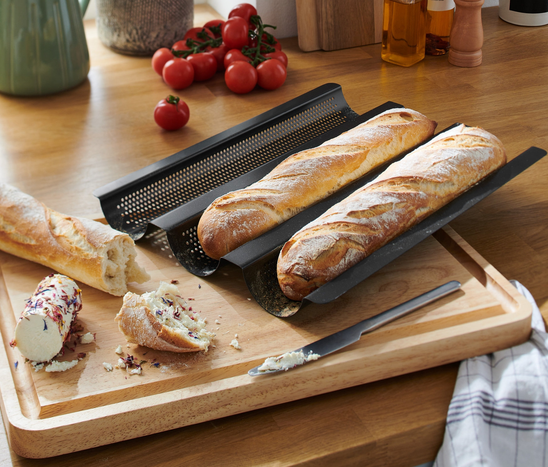 Dwie bagietki leżą w czarnej, perforowanej blasze do pieczenia bagietek na drewnianej desce do krojenia. Obok leży pokrojona bagietka z pastą do smarowania i nóż.
