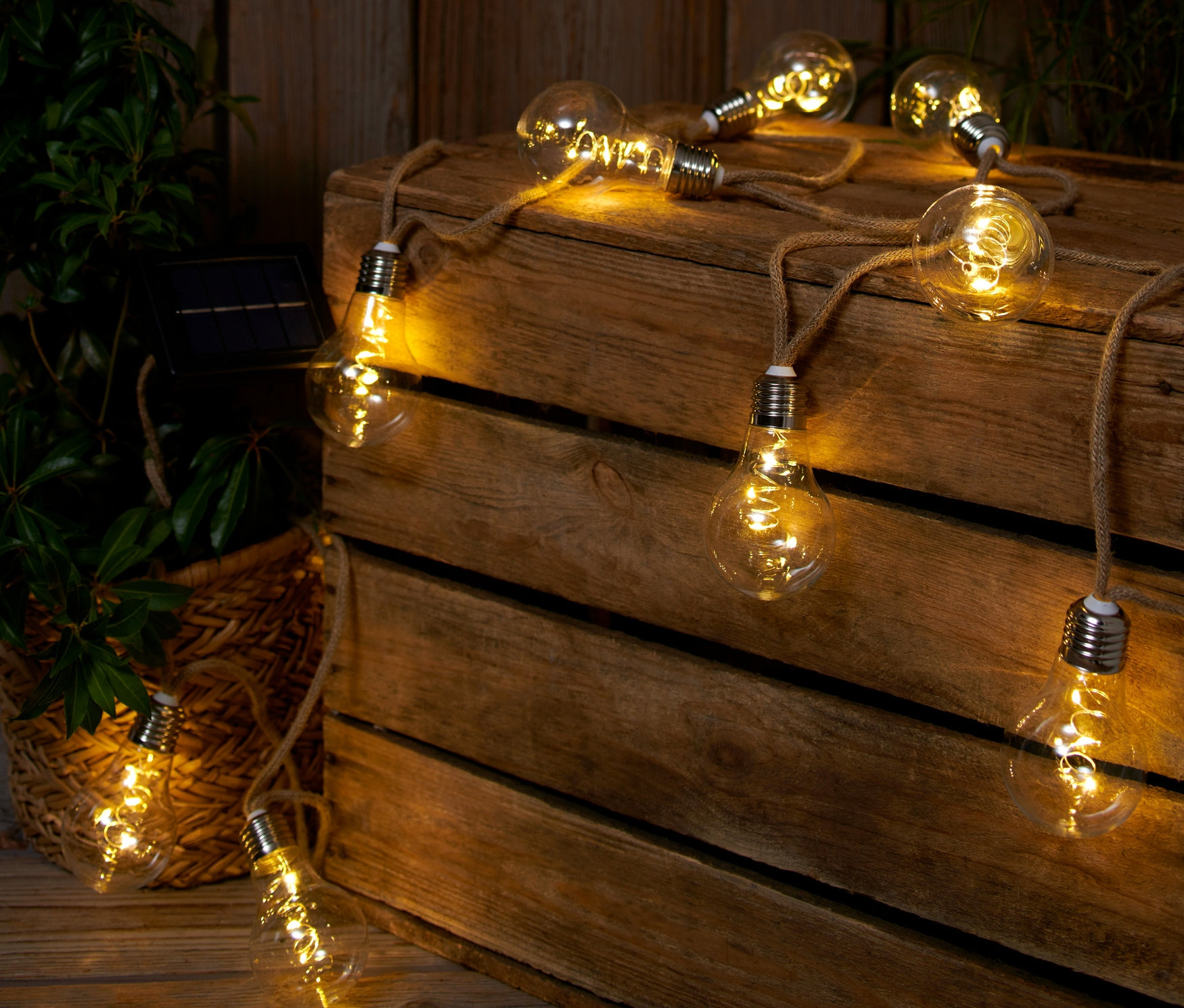 Solarny łańcuch świetlny z linką jutową leży na drewnianej skrzyni obok rośliny.