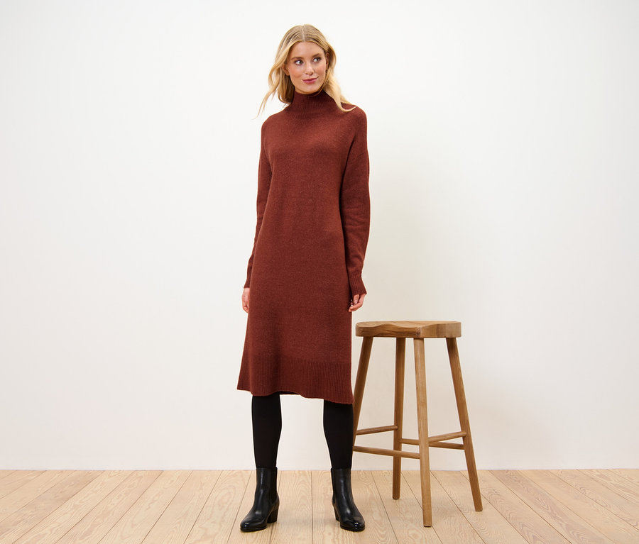 Kobieta stoi obok drewnianego stołka, ubrana w dzianinową sukienkę z zawartością wełny.