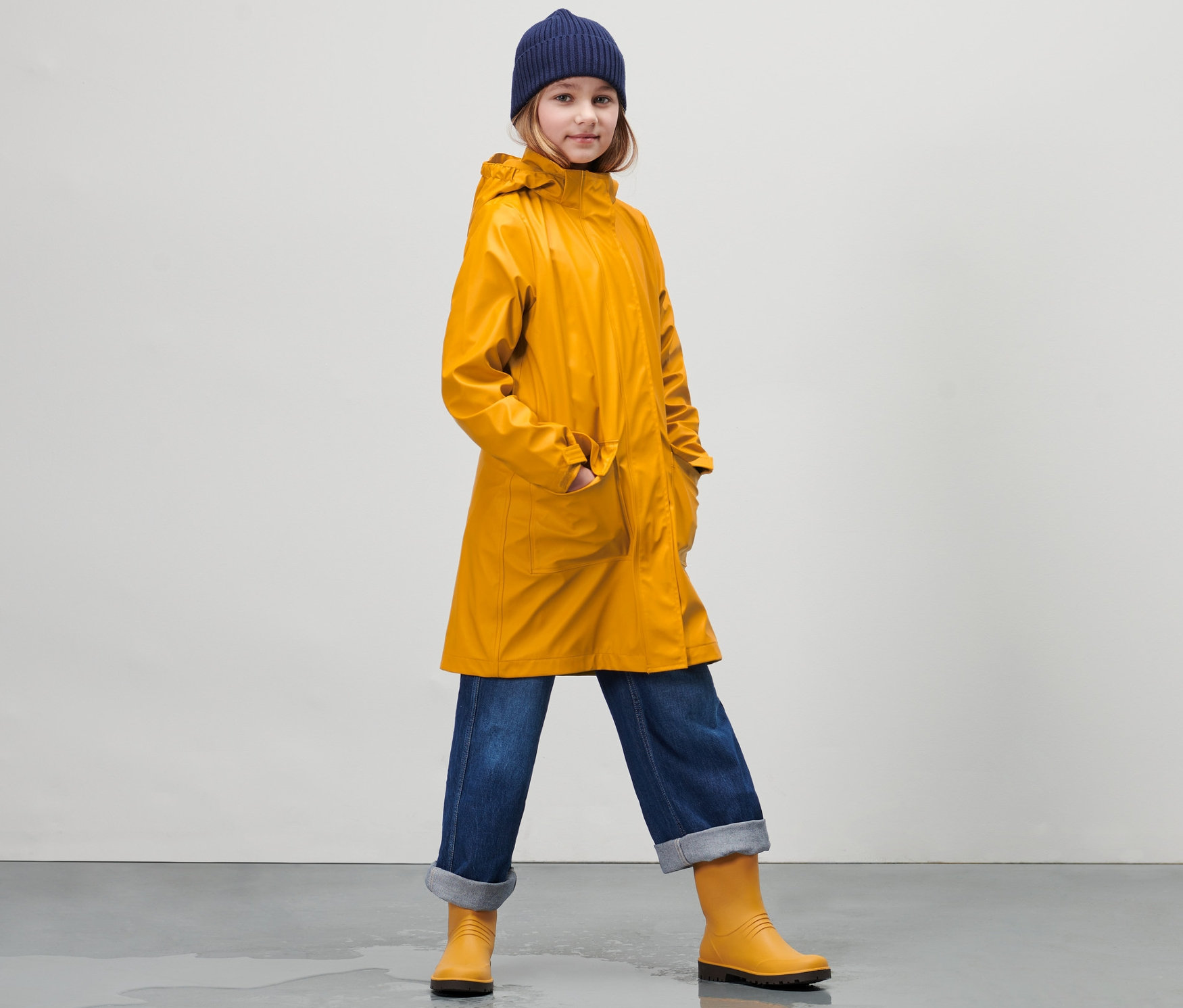 Dziewczyna w dziecięcej parce przeciwdeszczowej w stylu sztormiaka, dżinsach i kaloszach stoi na mokrej podłodze.