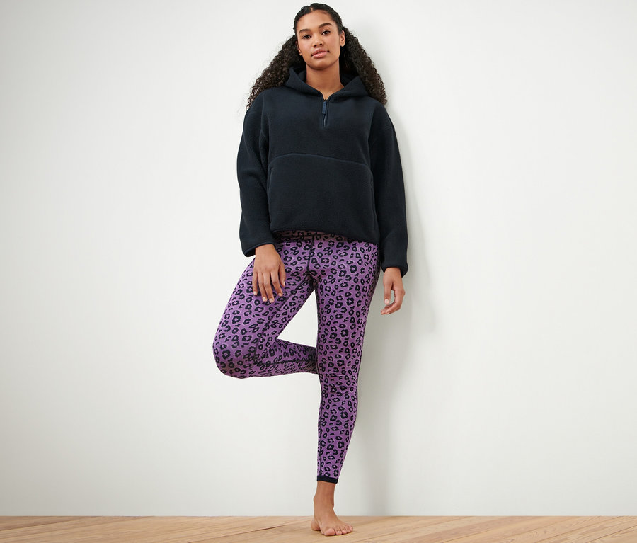Kobieta stoi na jednej nodze, ubrana w granatową bluzę z kapturem z pluszu i dwustronne legginsy sportowe, niebieskie i lawendowe.