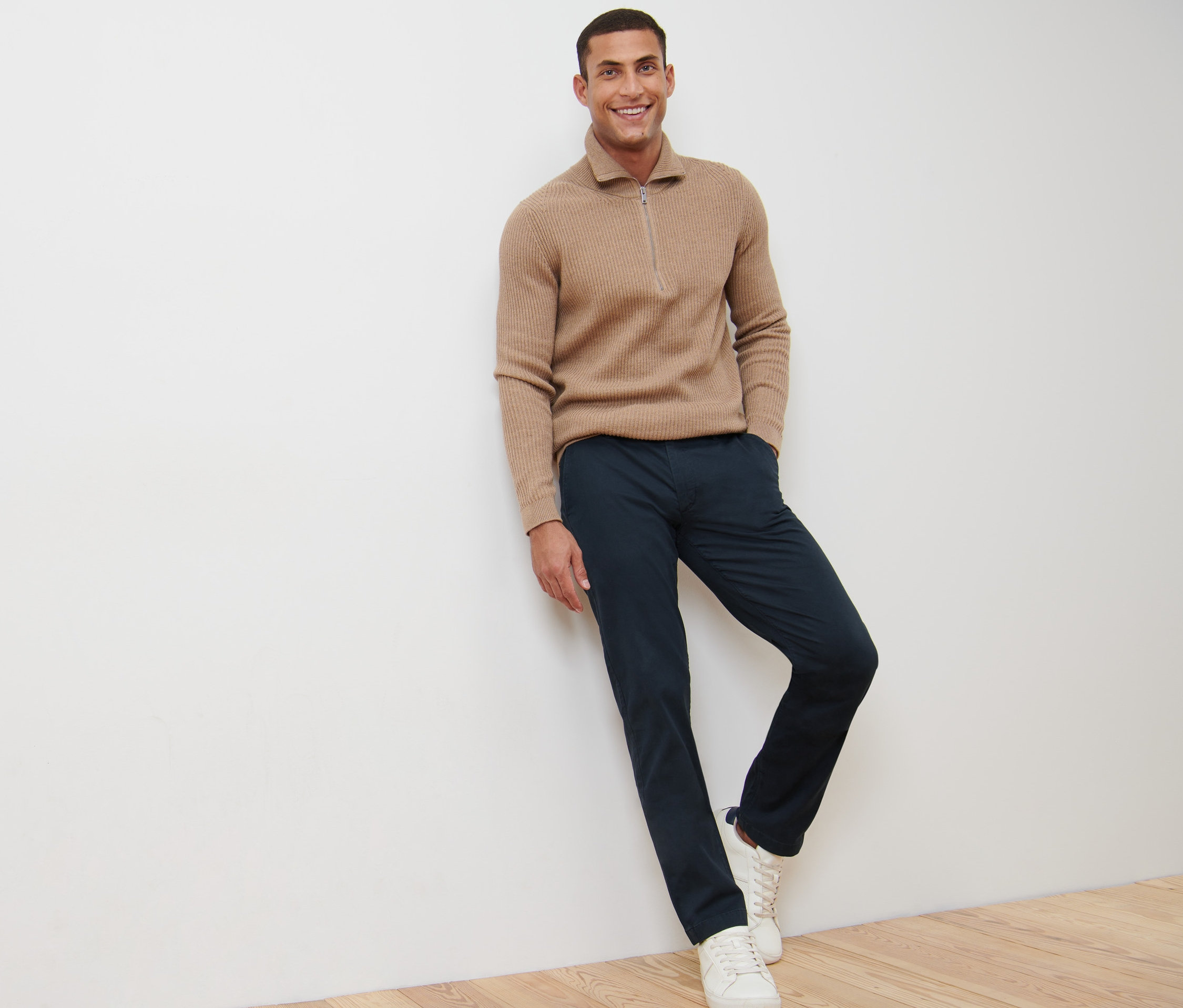 Uśmiechnięty mężczyzna opiera się o ścianę w granatowych spodniach chinosach.