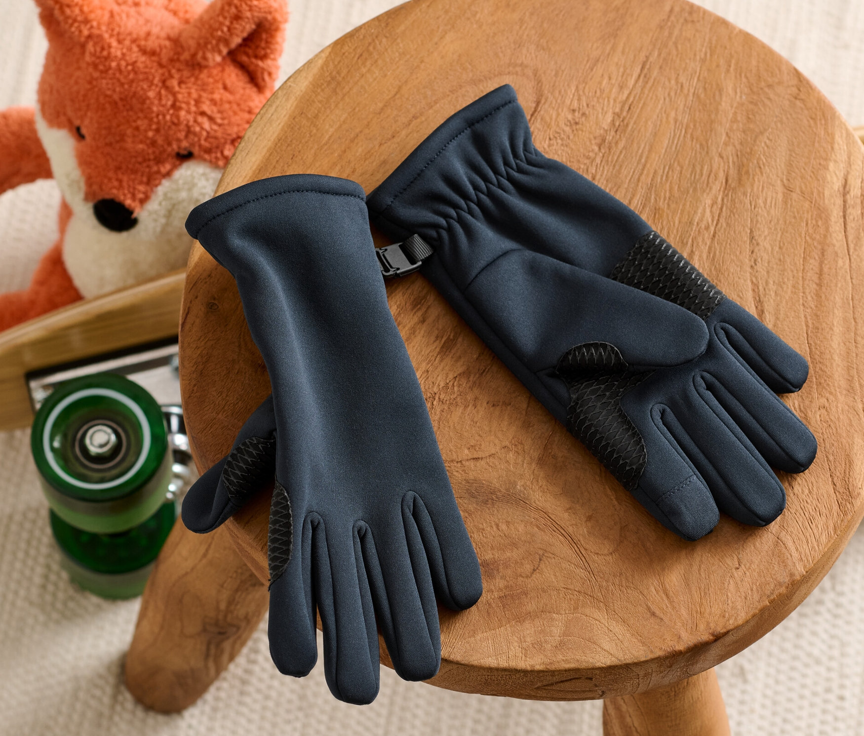 Rękawiczki z softshellu leżą na drewnianym stołku, w tle pluszowy lis na deskorolce.
