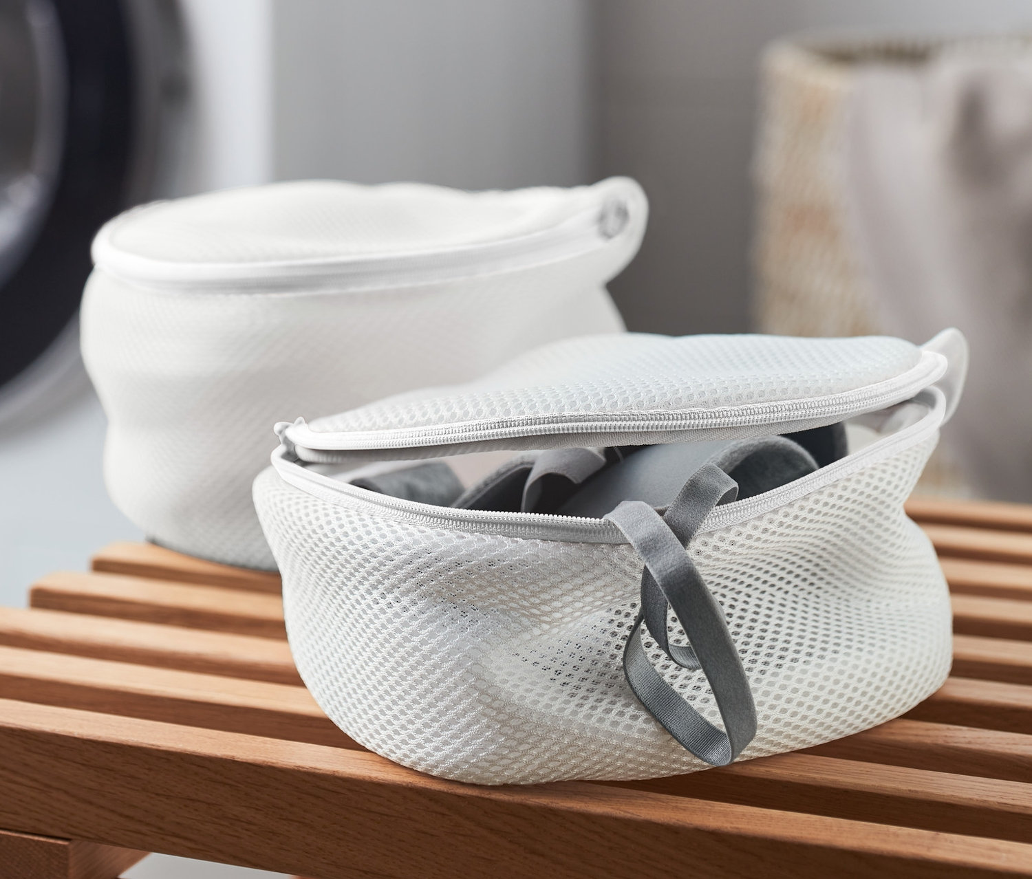 Dwa białe woreczki do prania biustonoszy na drewnianej ławce, w tle pralka.