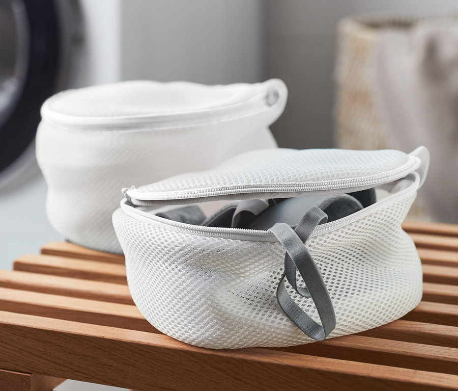 Dwa białe woreczki do prania biustonoszy na drewnianej ławce, w tle pralka.