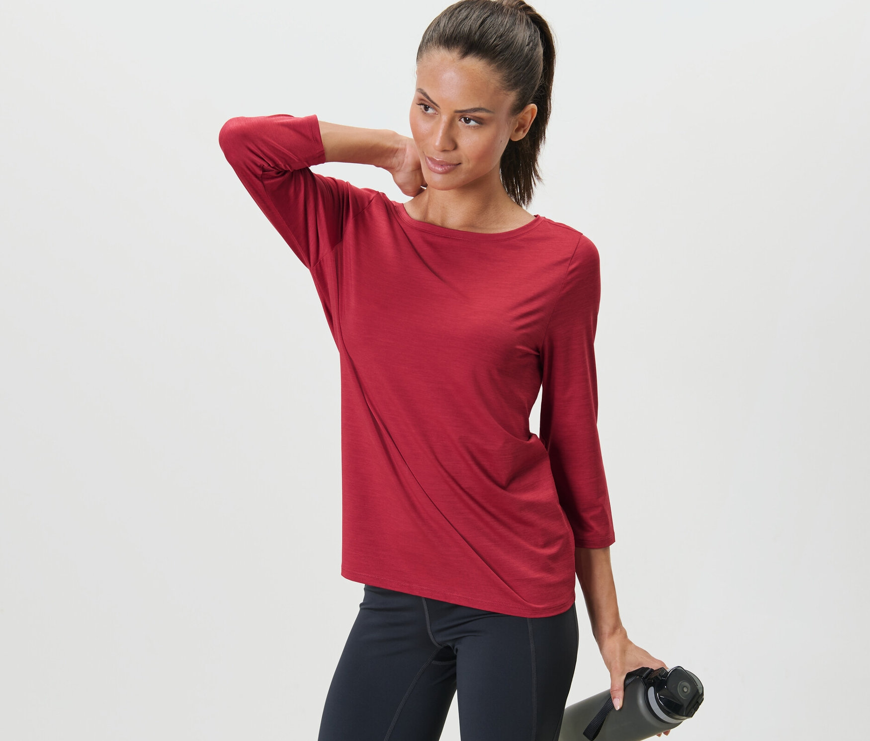 Kobieta pozuje w czerwonej koszulce funkcyjnej z rękawem 3/4 i czarnych legginsach, trzymając w ręku butelkę z wodą.