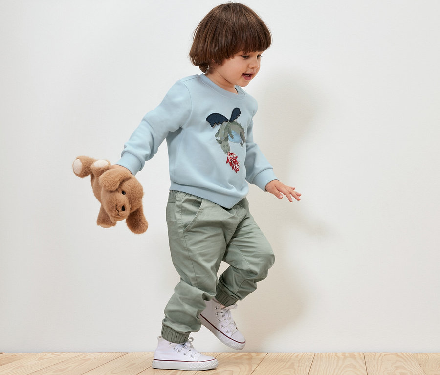 Chłopiec w niebieskiej bluzie z nadrukiem smoka i zielonych spodniach Spodnie na gumce – fason „Anton” trzyma pluszowego psa i idzie po drewnianej podłodze.