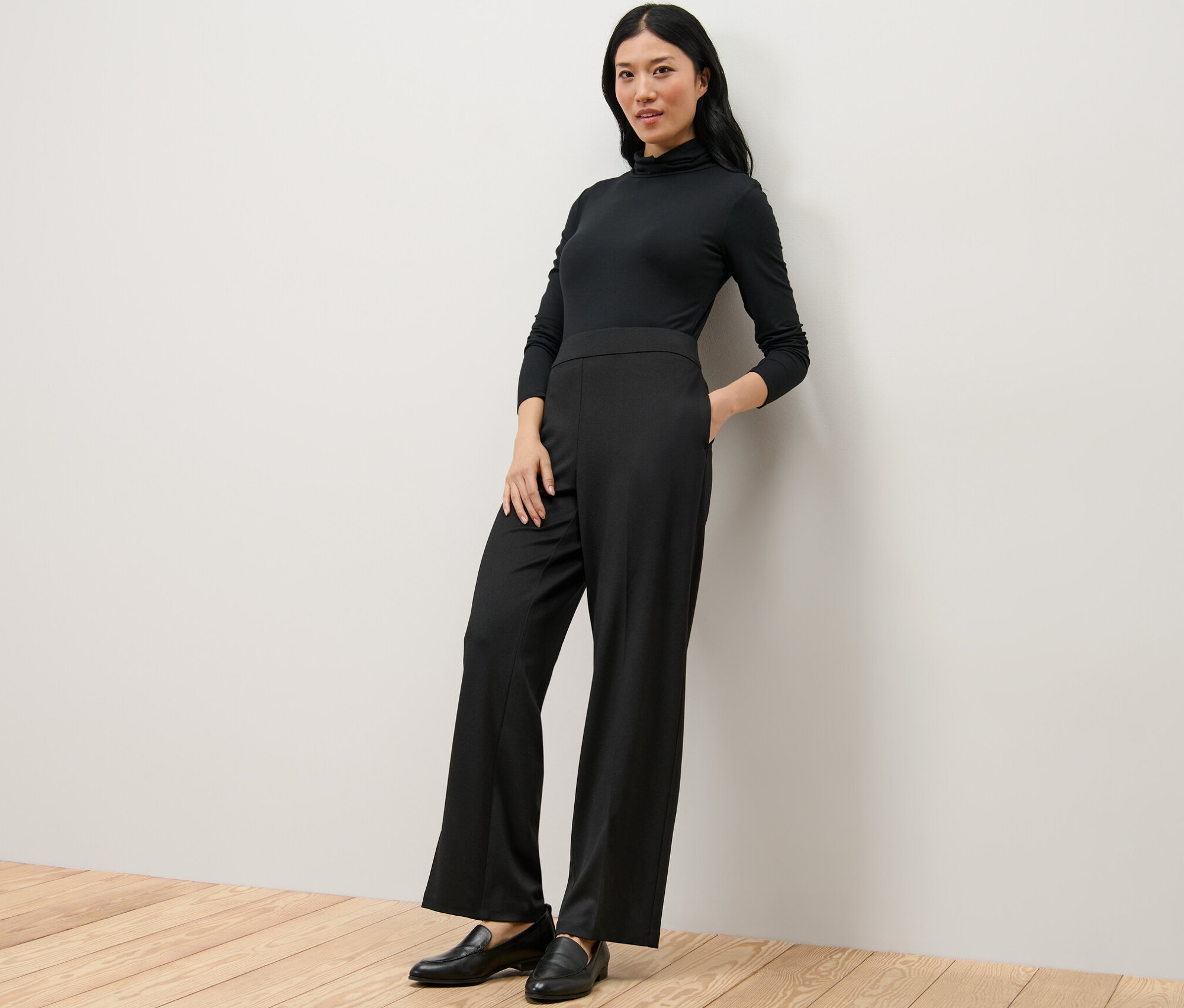 Kobieta w czarnym golfie i wygodnych spodniach z dżerseju opiera się o ścianę.