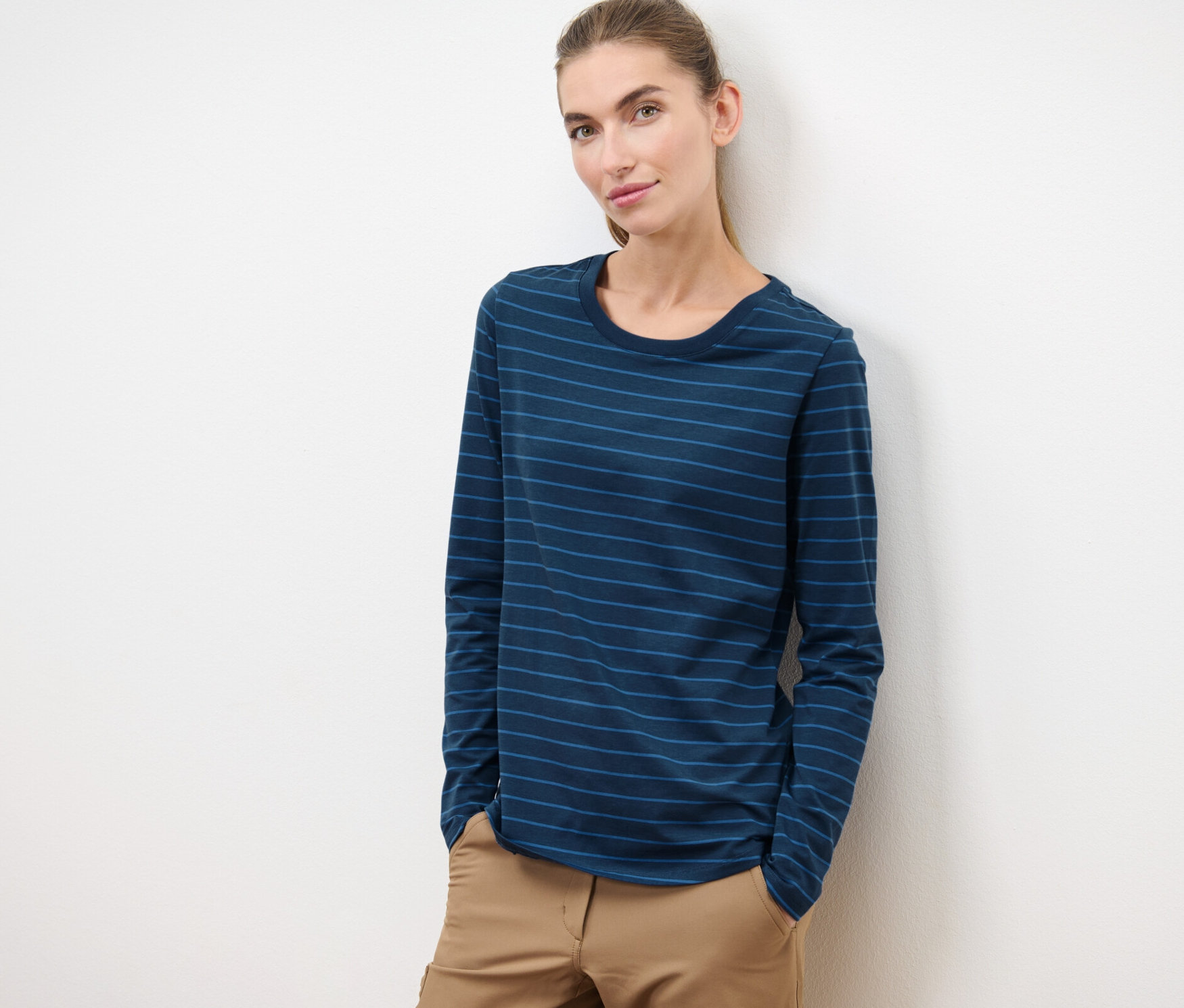 Kobieta opiera się o białą ścianę, ubrana w koszulkę sportową z długim rękawem i brązowe spodnie.