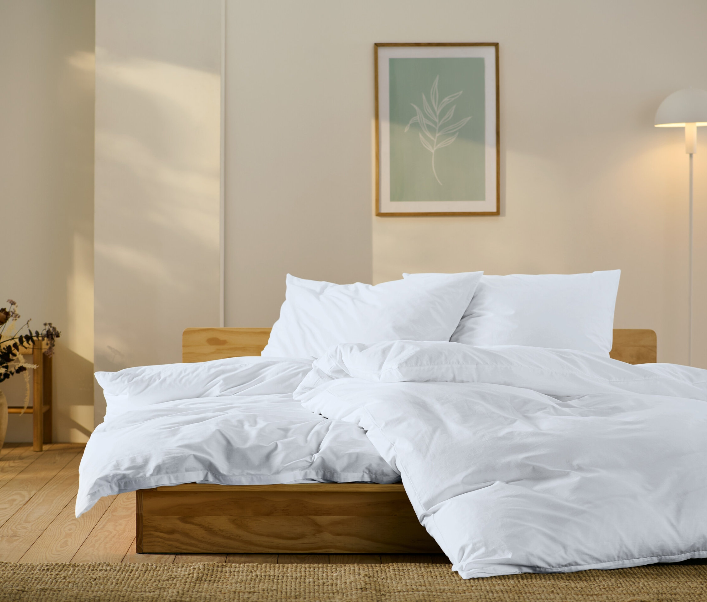 Drewniane łóżko z białą pościelą, poduszkami i kołdrą. Na ścianie obraz z motywem liścia i lampa.