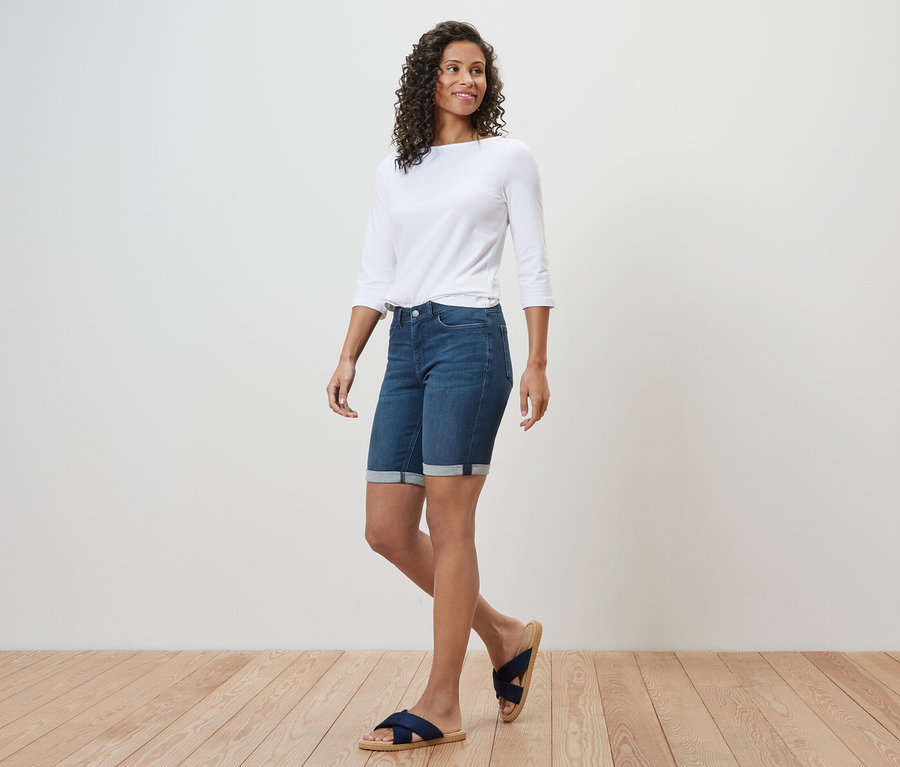 Kobieta idzie, ma na sobie dżinsy bermudy – fason „Lea”, dark blue denim, biały top z rękawami trzy czwarte i sandały.