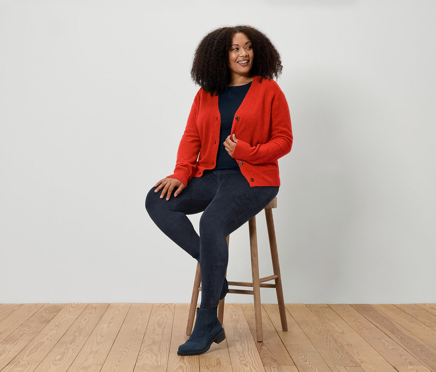 Kobieta siedzi na stołku, ubrana w czerwony kardigan, niebieskie dżinsy i niebieskie buty.