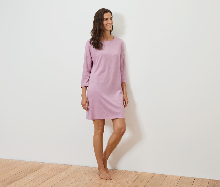 Kobieta stoi i uśmiecha się, ma na sobie fioletową sukienkę z rękawami trzy czwarte.