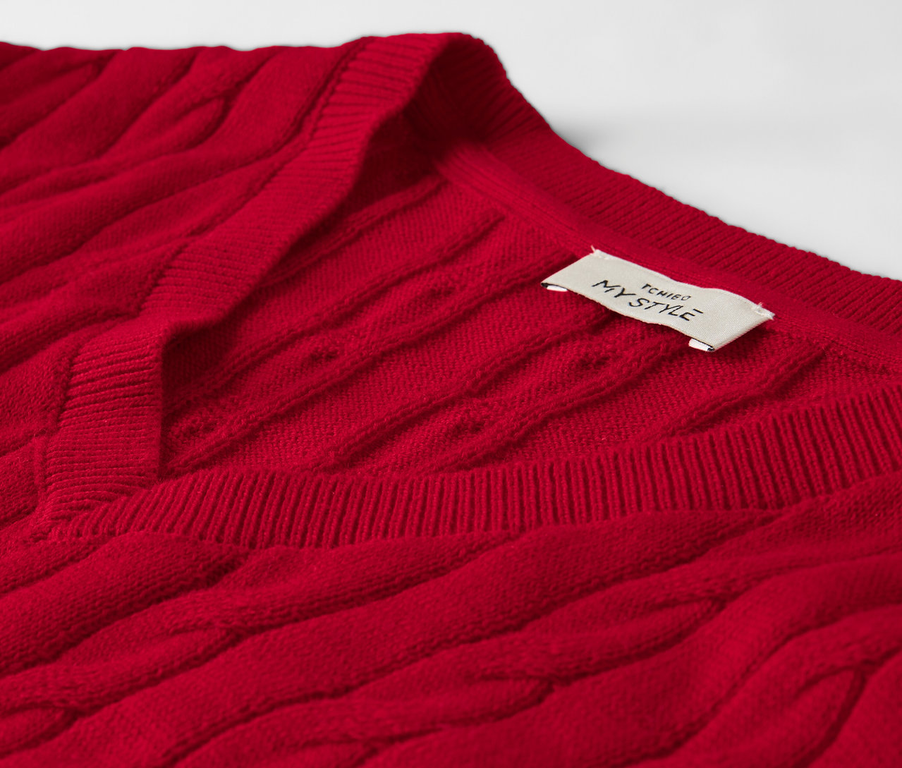 Zbliżenie na czerwony sweter w warkocze z metką przy dekolcie.