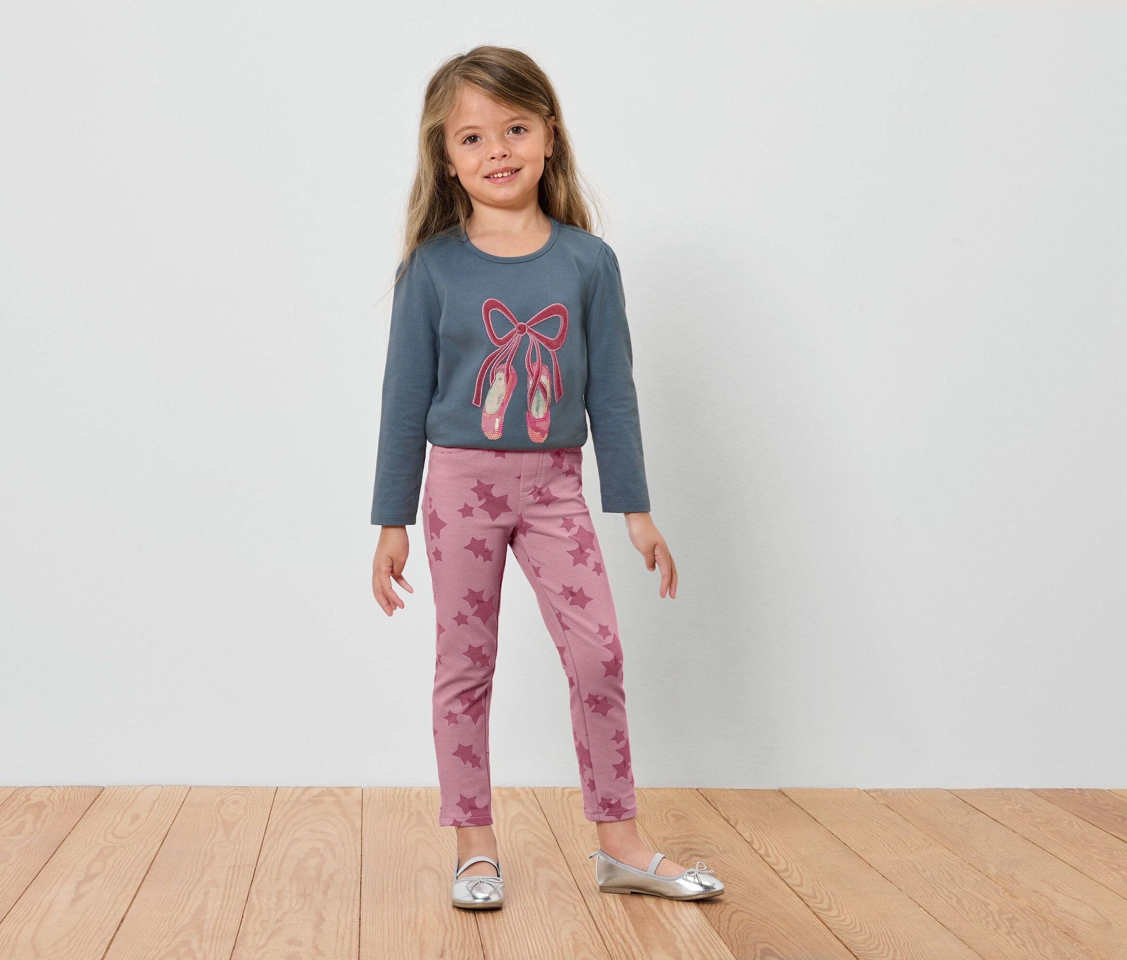 Dziewczynka w niebieskiej bluzce z długim rękawem z baletkami i różowych legginsach stoi na drewnianej podłodze.