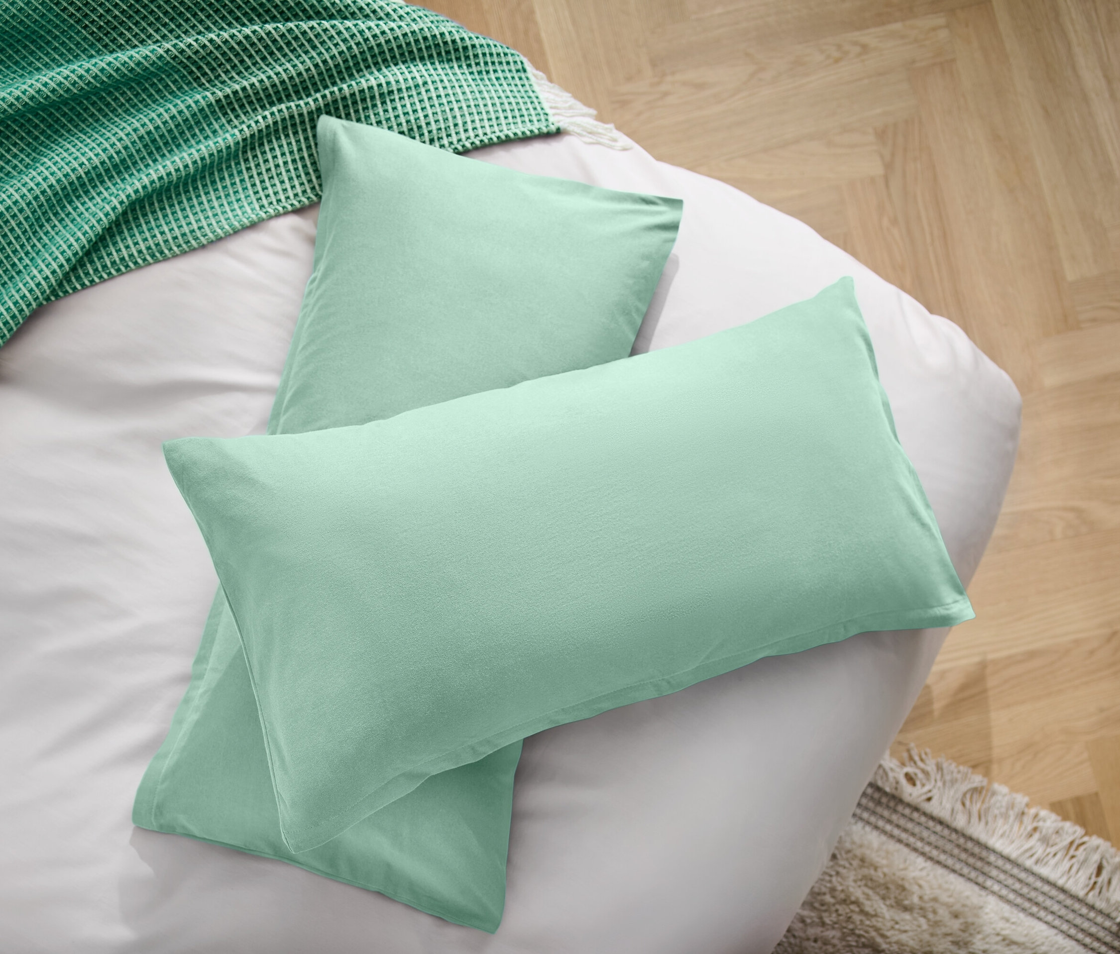 Dwie jasnozielone poduszki na białym prześcieradle obok zielonego koca.