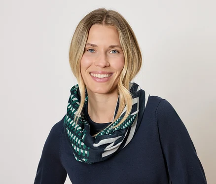 Kobieta z blond włosami i uśmiechem ma na sobie granatowy sweter i szalik z geometrycznym wzorem w kolorze zielonym i białym.