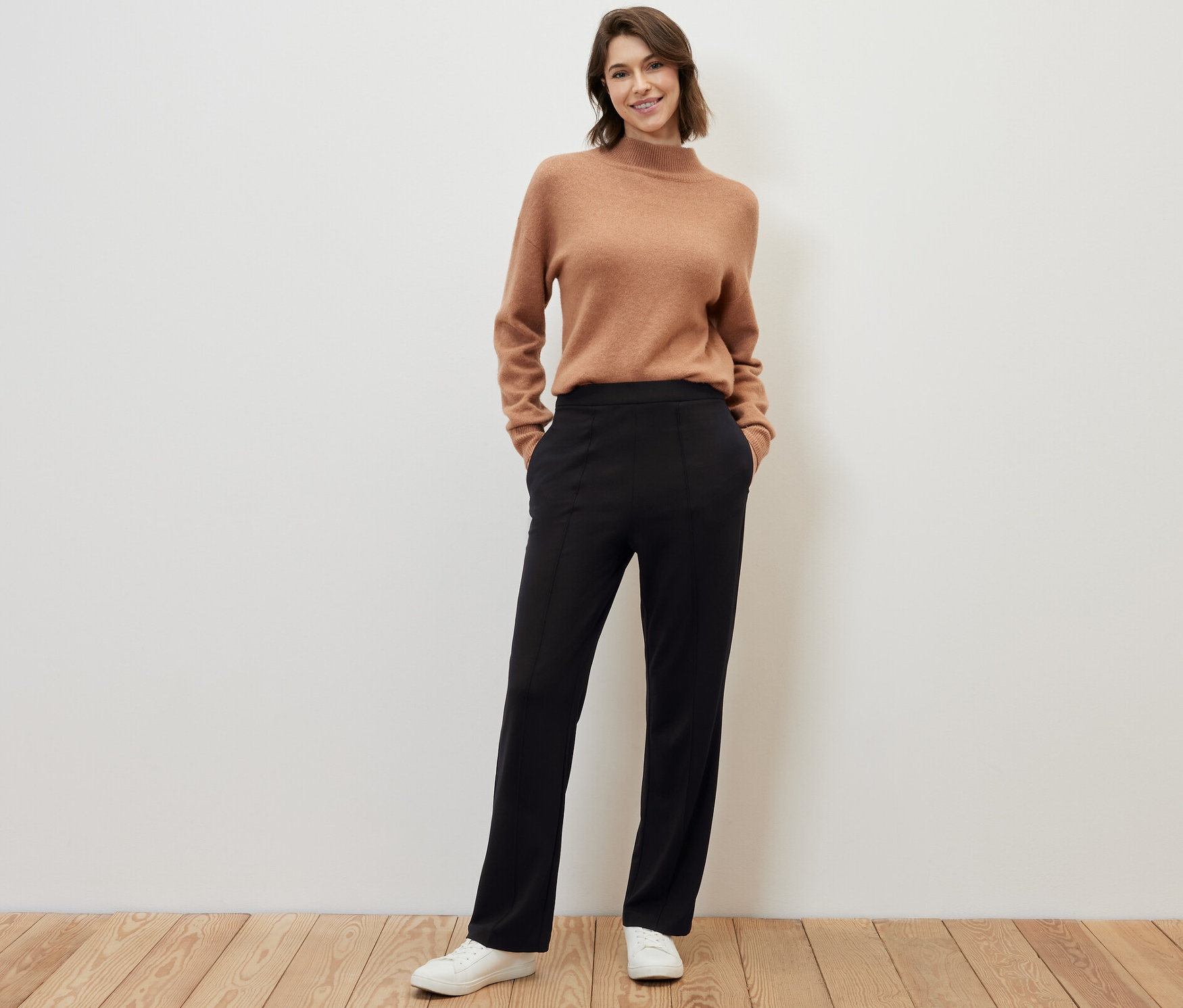 Kobieta pozuje na drewnianej podłodze w brązowym swetrze i czarnych spodniach dżersejowych z kantem.