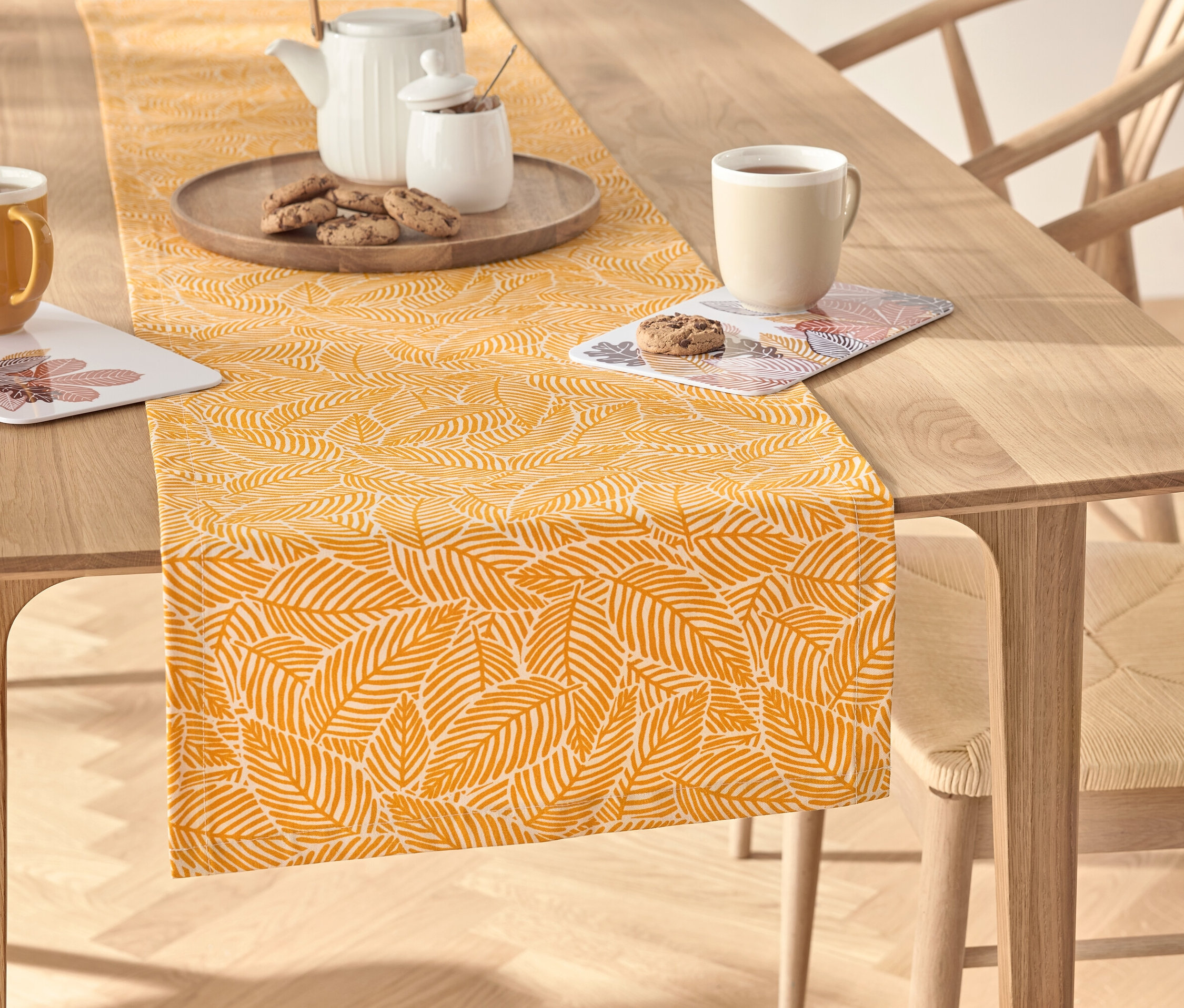Na drewnianym stole leży żółty bieżnik ze wzorem liści, na którym znajduje się serwis do herbaty, filiżanka i ciasteczka.