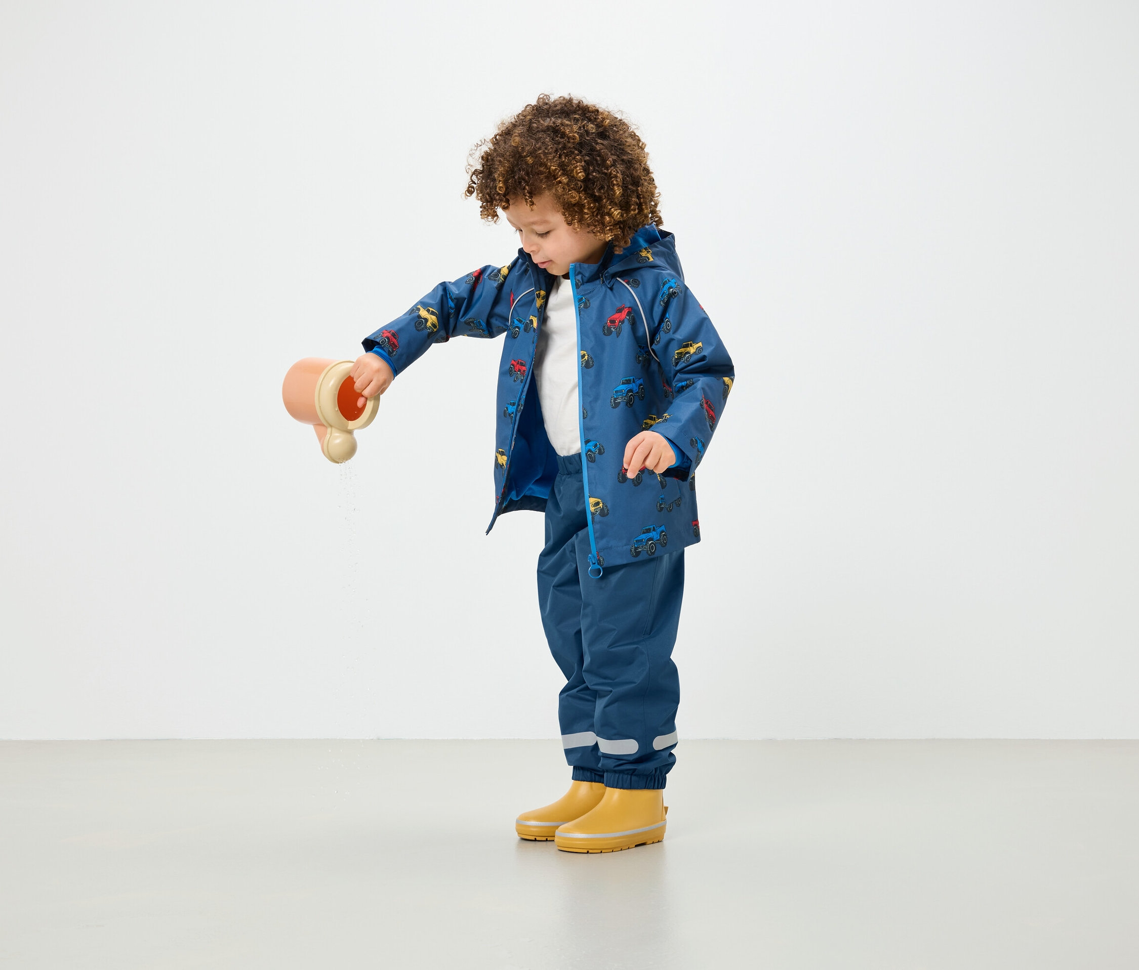 Dziecko z kręconymi włosami wylewa wodę z zabawki, ubrane w niebieski płaszcz przeciwdeszczowy w nadruki i niebieskie spodnie.