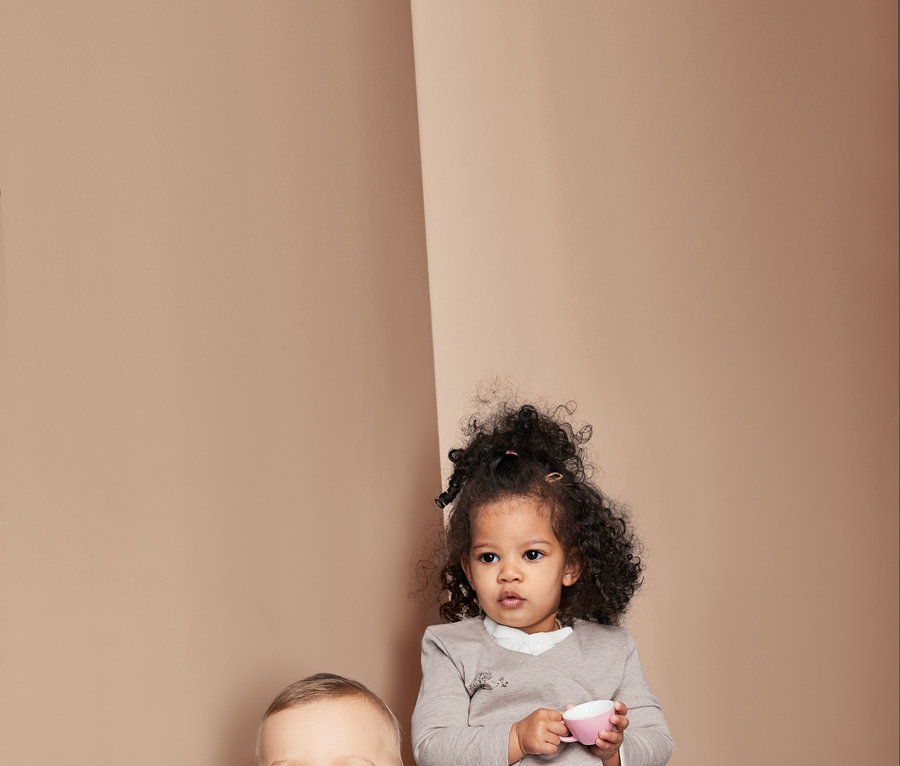 Dwoje dzieci siedzi. Chłopiec siedzi na poduszce i uśmiecha się, ma na sobie koszulkę w paski. Dziewczynka trzyma różowy kubek i ma na sobie top. Oboje mają na sobie dżinsy.