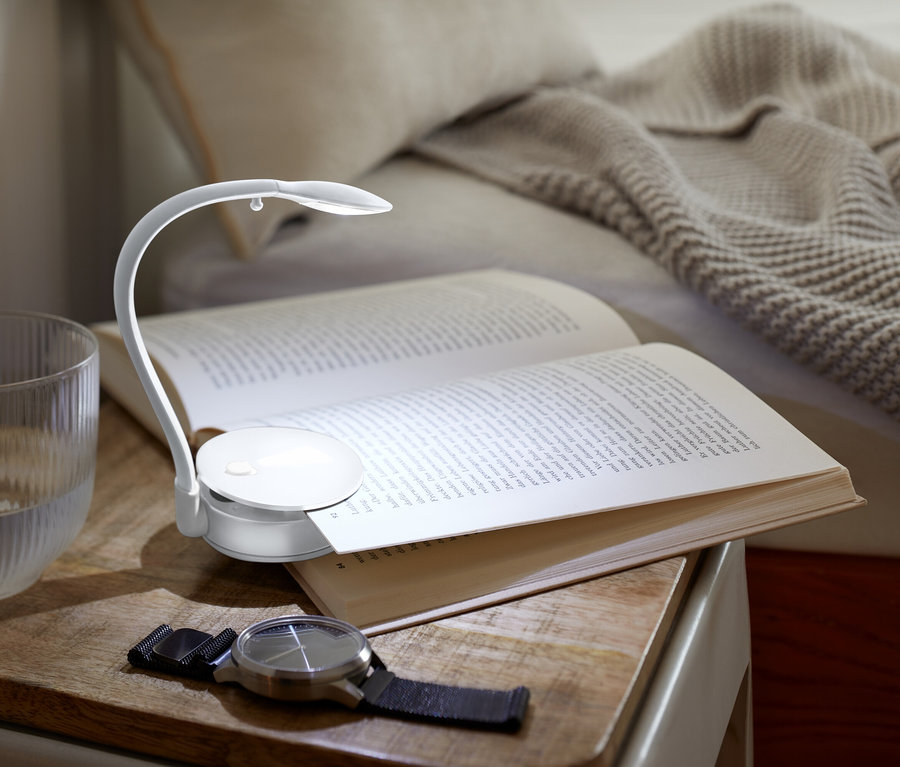 Na stoliku nocnym leży otwarta książka oświetlona małą lampką, obok stoi szklanka wody i zegarek.