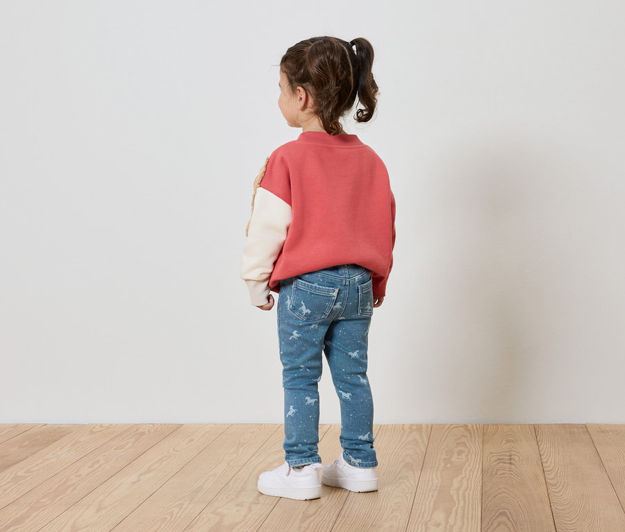 Dziewczynka w różowej bluzie, niebieskich dżinsach z nadrukiem i białych butach stoi tyłem do kamery.
