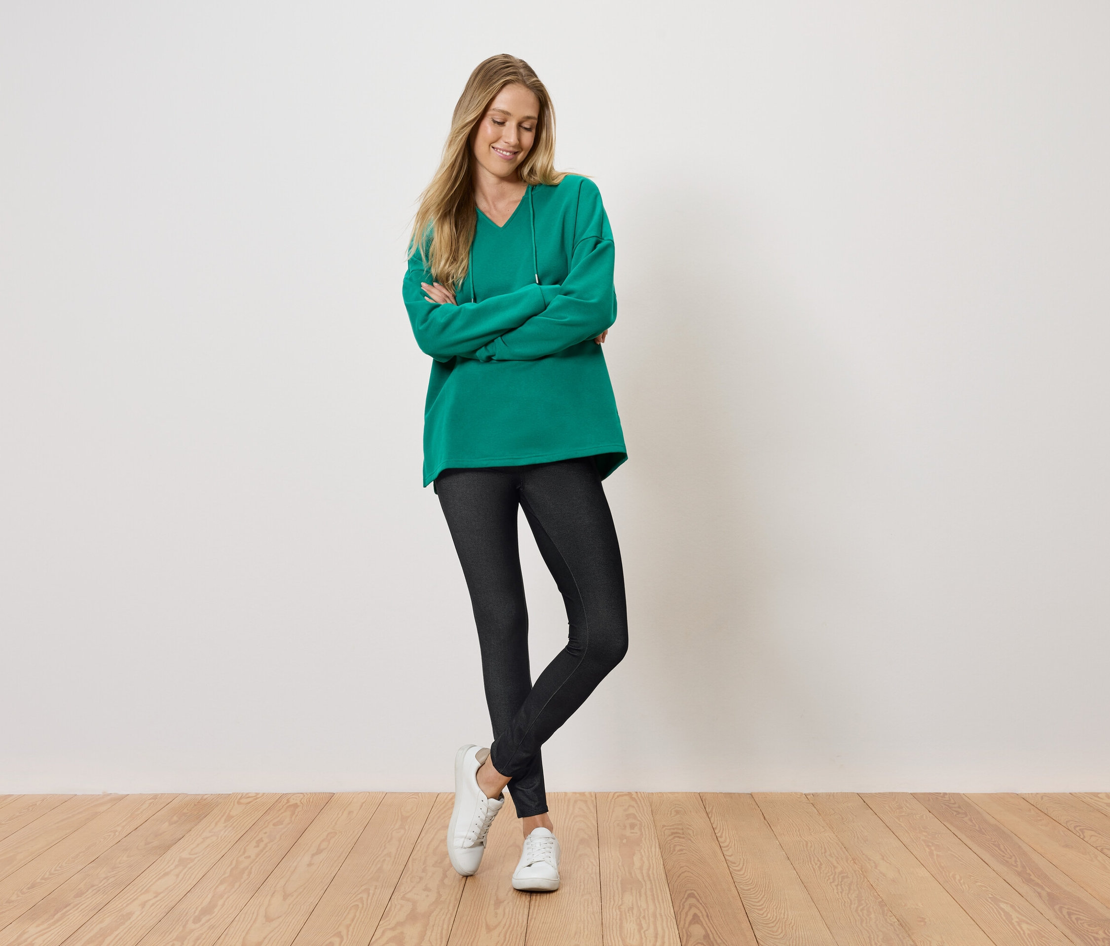 Kobieta stoi z założonymi rękami i uśmiecha się, ubrana w zielony sweter, czarne legginsy i białe buty.