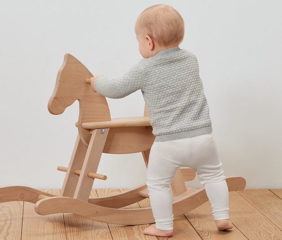 Niemowlę w rozpinanym swetrze opiera się o drewnianego konika na biegunach.