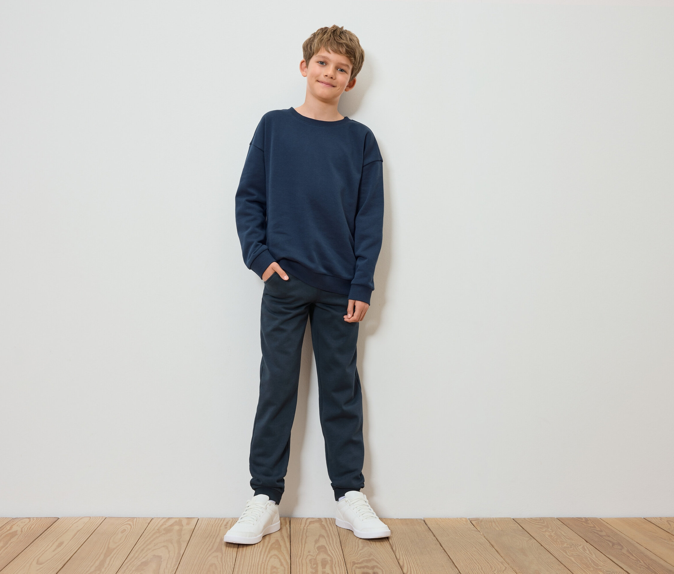 Chłopiec stoi i pozuje w granatowej bluzie, spodniach i białych tenisówkach.