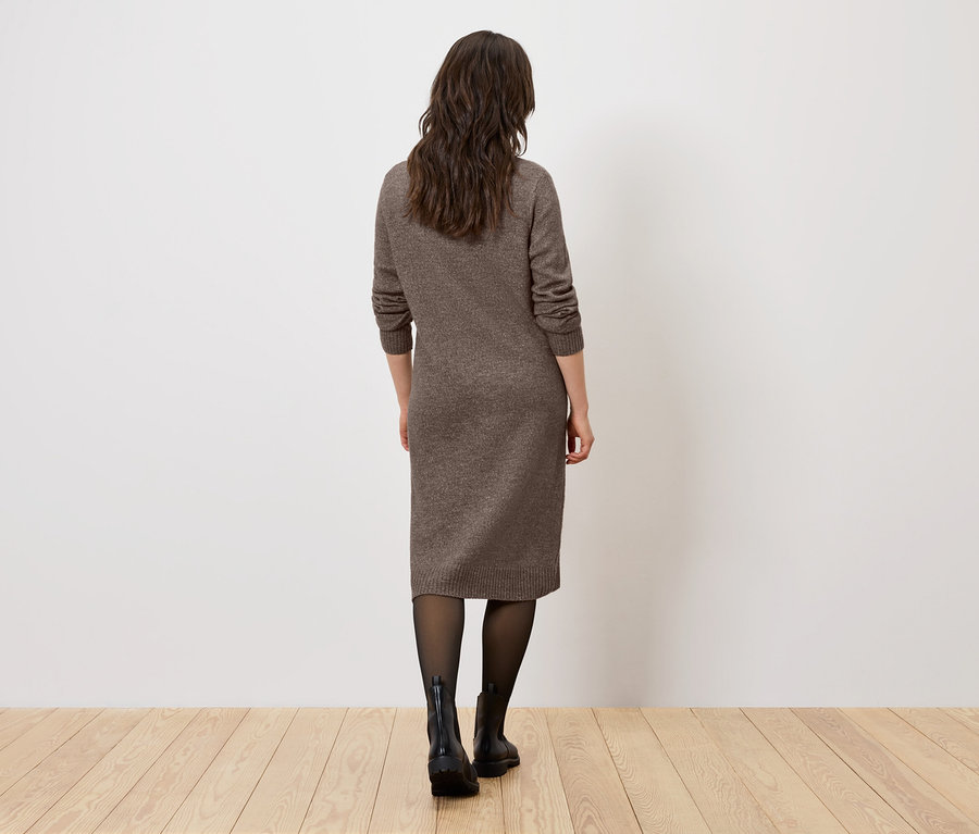 Kobieta tyłem do kamery stoi na drewnianej podłodze, ubrana w brązową sukienkę, czarne rajstopy i czarne buty.