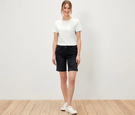 Kobieta stojąca na drewnianej podłodze ma na sobie dżinsy bermudy – fason „Lea”, black denim i białą koszulkę.