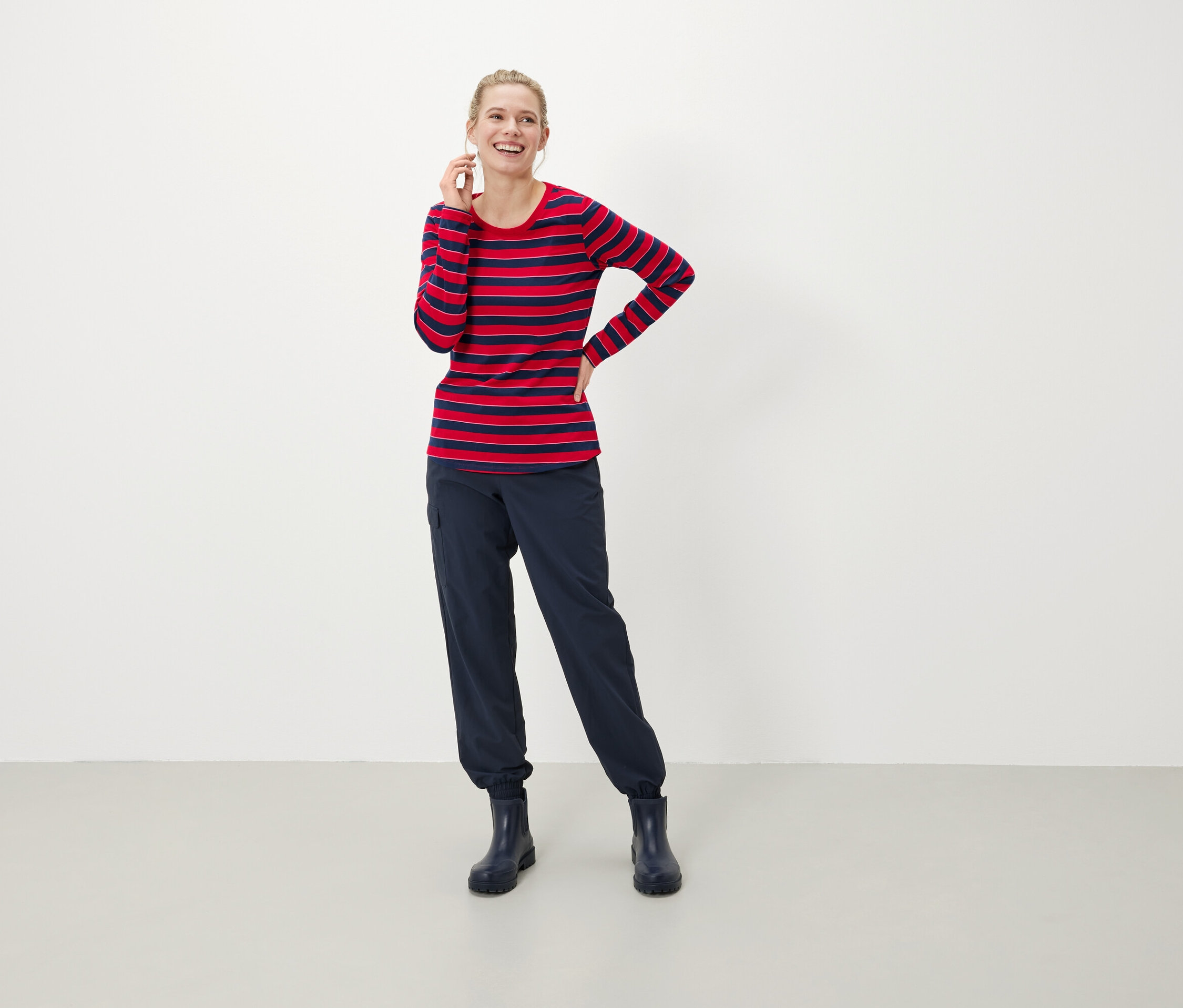 Kobieta z blond włosami uśmiecha się i trzyma telefon przy uchu. Ma na sobie czerwono-niebieską bluzkę w paski z długim rękawem, granatowe spodnie i buty.