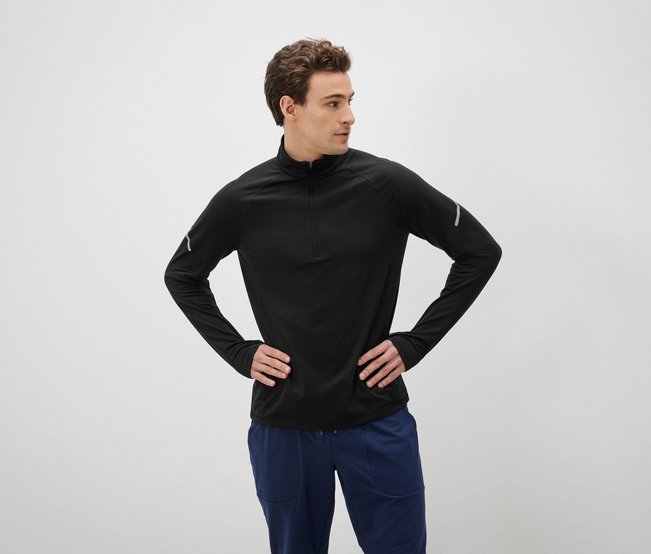 Mężczyzna z brązowymi włosami pozuje z rękami na biodrach, ubrany w czarną koszulkę z długim rękawem i niebieskie spodnie.