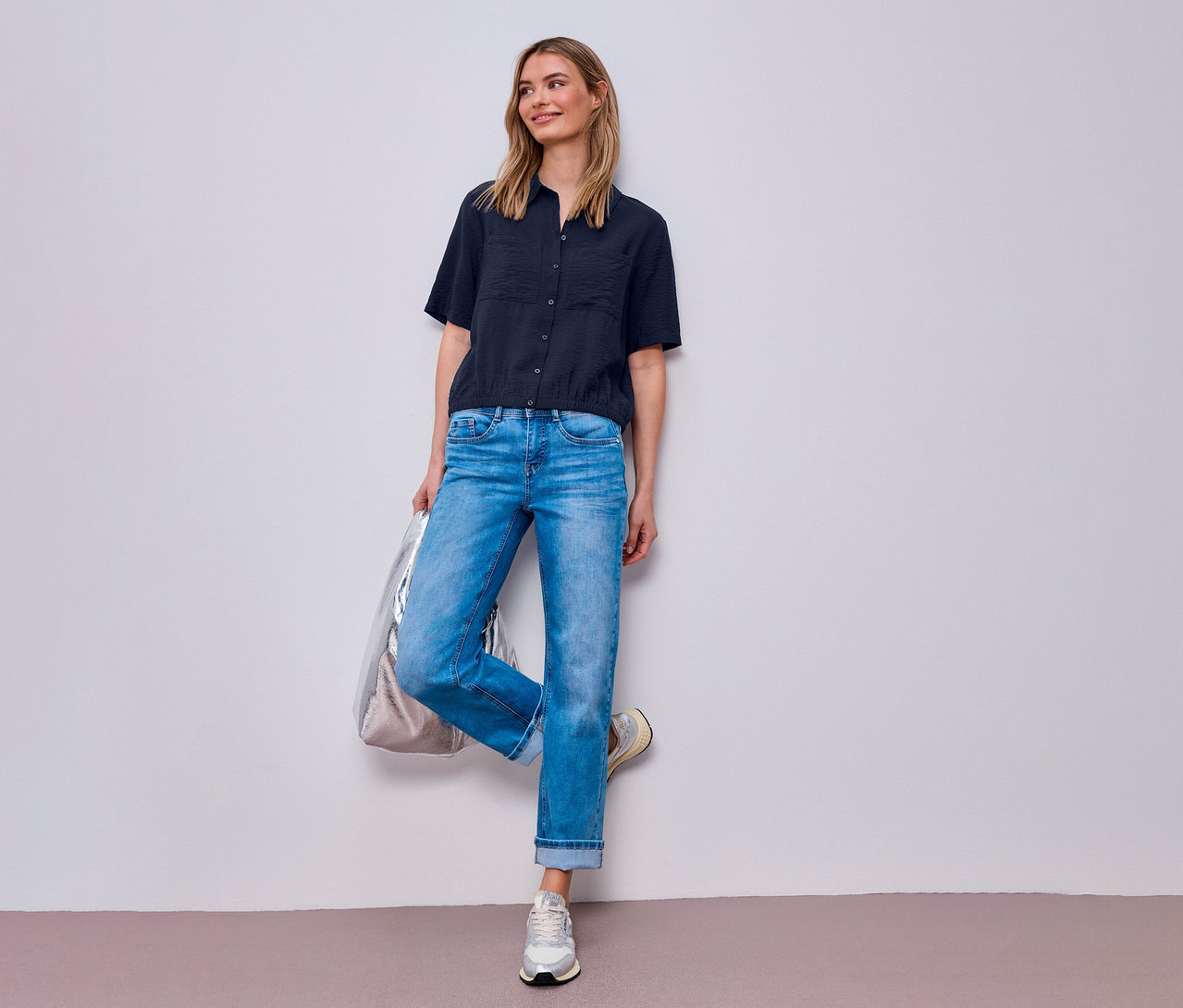 Kobieta w dżinsach i koszuli Street One pozuje ze srebrną torbą przy ścianie.