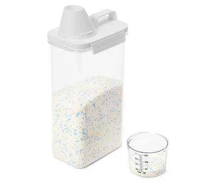 Przezroczysty pojemnik z białą pokrywką wypełniony jest detergentem do prania. Obok stoi miarka, która również zawiera detergent.