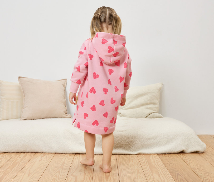 Dziewczynka stoi tyłem do kamery, ubrana w różową sukienkę z kapturem w serduszka.