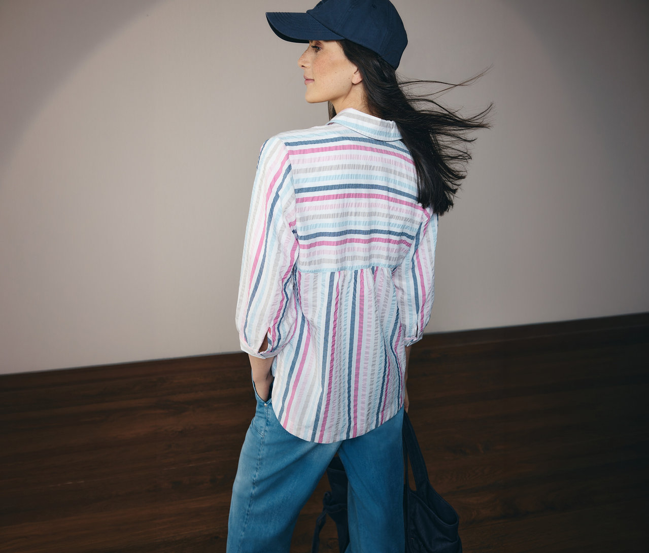Kobieta w dżinsach i bluzce koszulowej CECIL w niebieskie paski oraz czapce, widziana z tyłu.