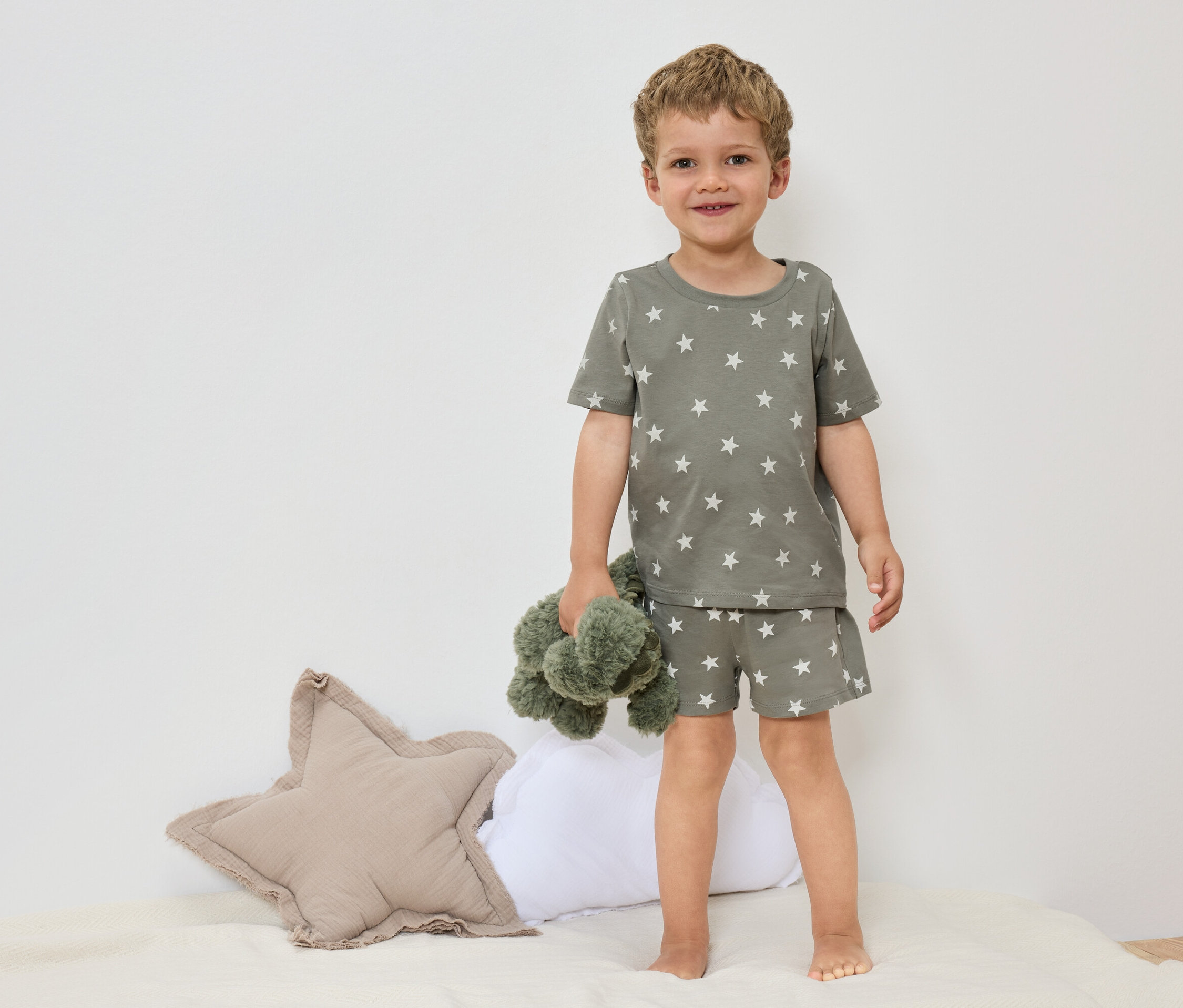 Mały chłopiec stoi boso na łóżku w oliwkowozielonej piżamie w gwiazdki, trzymając zieloną pluszową zabawkę.