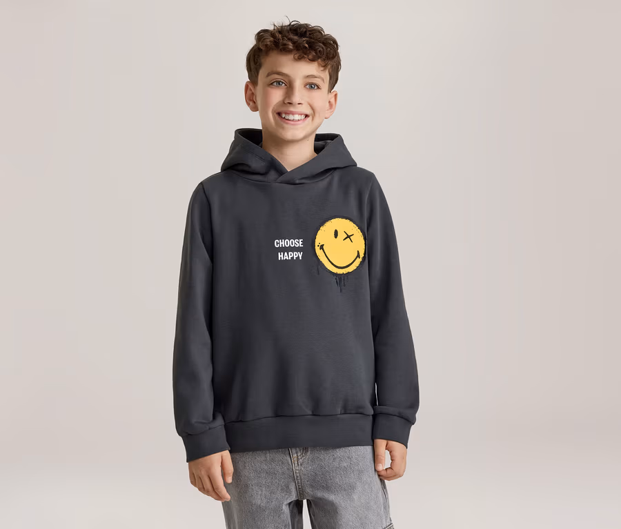 Uśmiechnięty chłopiec w ciemnoszarej bluzie z kapturem z żółtą buźką, która ma X zamiast oka i efekt topnienia, oraz napisem „CHOOSE HAPPY”. Ma na sobie jasnoszare dżinsy.