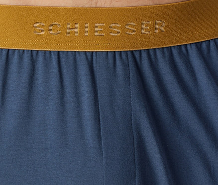 Zbliżenie niebieskiego materiału ze złotym elastycznym pasem, na którym widnieje nazwa marki „SCHIESSER” w perforowanych literach.